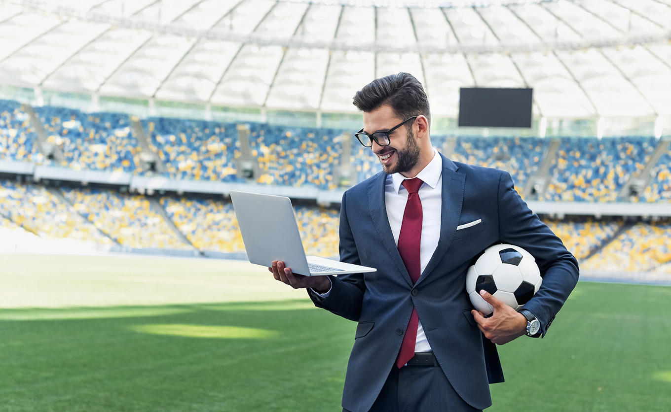 Fußballmanager mit Laptop in der Hand und Fußball unterm Arm im Fußballstadion