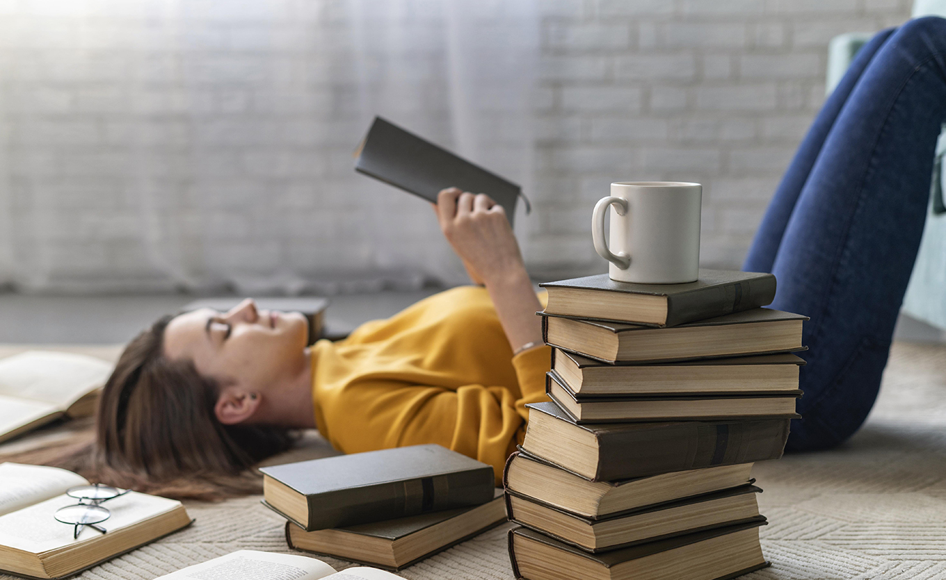 Frau liegt auf dem Boden zwischen einem Bücherstapel mit einem Kaffeebecher oben drauf und liest ein Buch